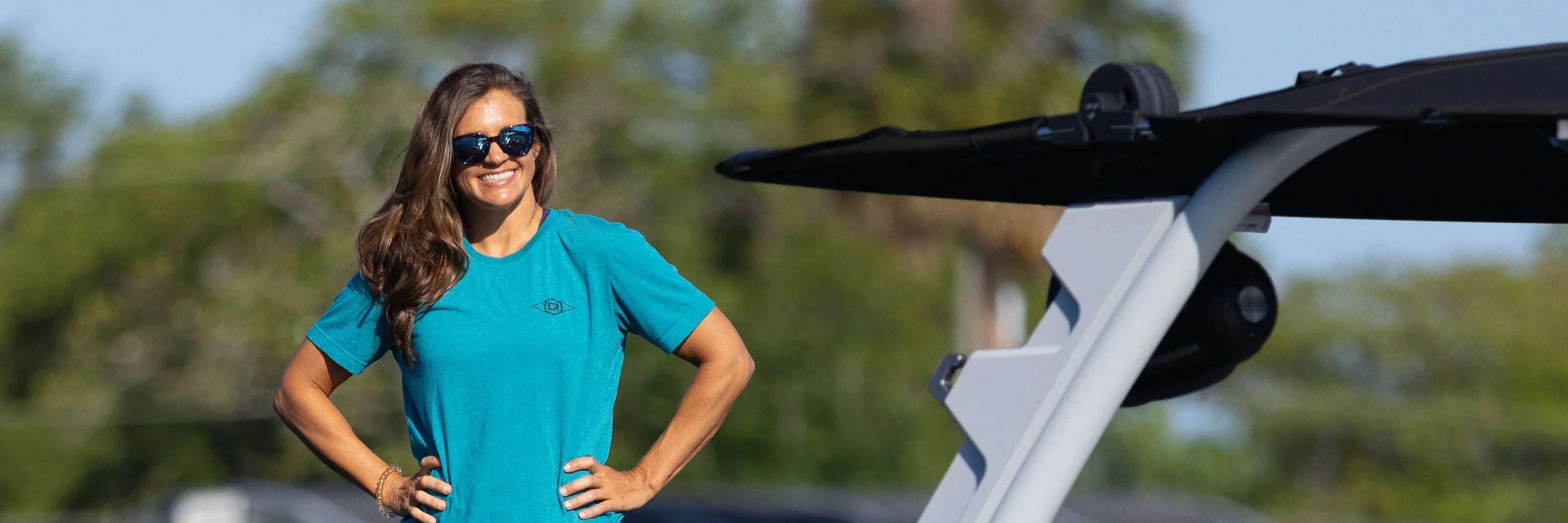 A woman wearing sunglasses and a teal O’Brien t-shirt stands next to a wakeboard boat with a confident smile.
