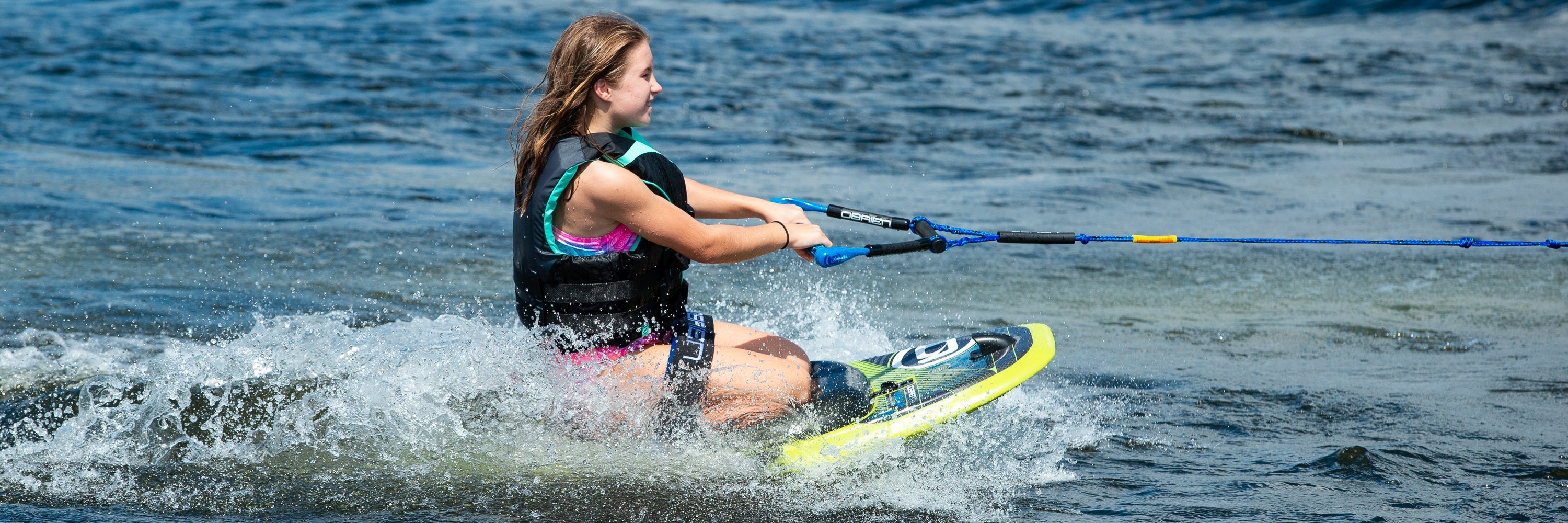 A girl in a black life vest and pink swimsuit skims across a lake on a kneeboard.