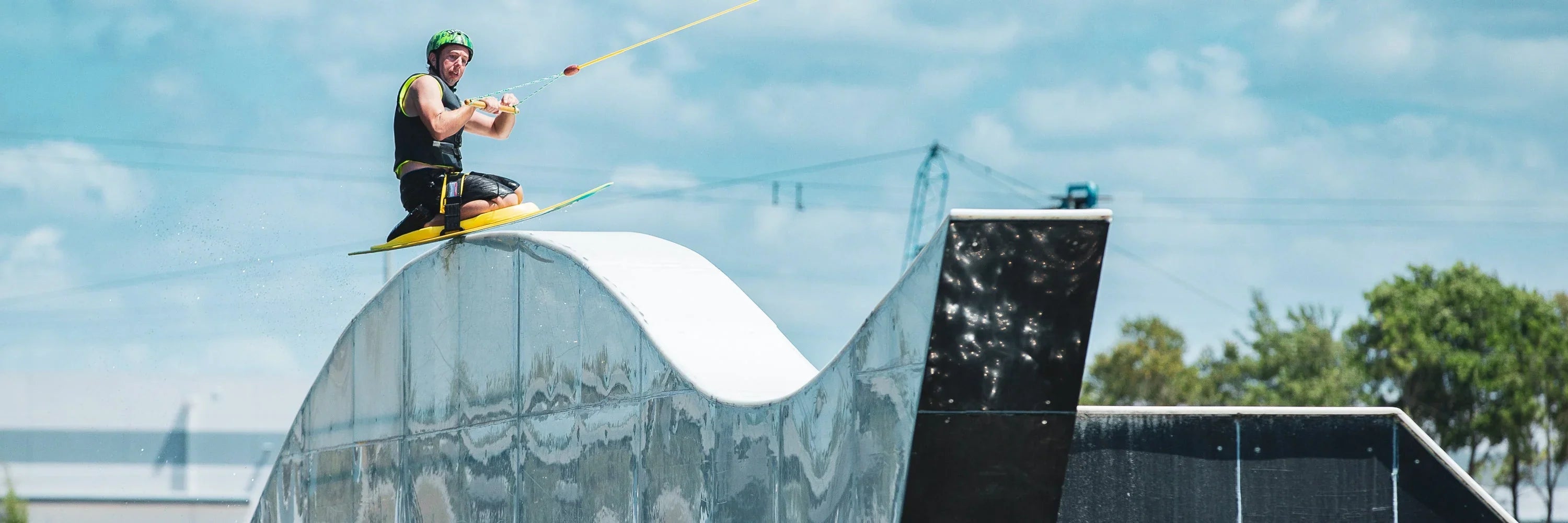 A kneeboarder in a helmet and vest rides up a white ramp at a cable park.