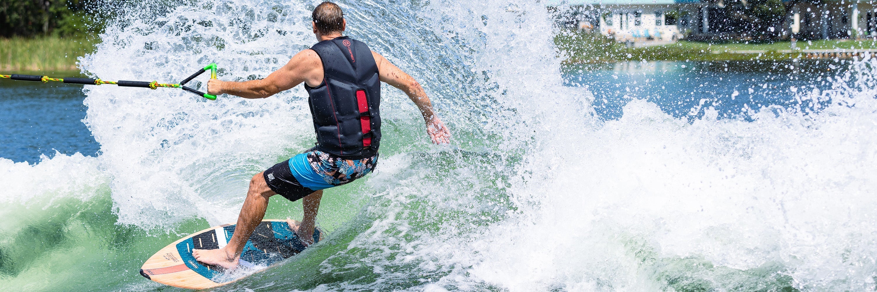 A man in a black vest riding the O'Brien Havana Wakesurf Board, cutting through a large wave with a powerful stance.